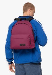 Bordový batoh s hladkým povrchem, přední kapsa na zip a nastavitelnými černými popruhy. Plochá nášivka s logem na přední straně. Nosen na modrém tričku.