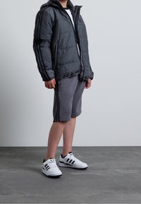 Puffa bunda v tmavě šedé barvě, s vertikálními černými pruhy, šedé šortky a bílé tenisky s černými akcenty a třemi pruhy.