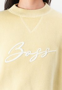 Vaaleankeltainen neulepaita, jossa on ribbed-tyylinen pyöreä pääntie ja valkoinen, kursiivilla kirjoitettu "BOSS" -logo edessä. Kankaan tekstuuritiedot näkyvät.