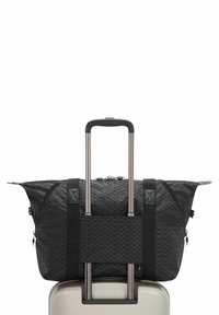 Un sac de sport noir à motifs fixé sur la poignée d'une valise rigide beige à roulettes avec une poignée télescopique déployée.