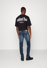 Camiseta gráfica negra con texto blanco en la parte posterior, combinada con jeans de mezclilla azul oscuro y zapatillas negras, vista desde atrás.