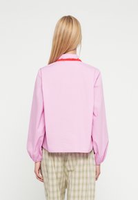 Roosa pluus pikkade punnitavate varrukatega ja punase dekoratiivse äärisega krae ümber, kombineeritud rohevalgete ruuduliste pükstega. Sile kangas.