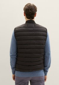 Schwarze Pufferweste mit horizontalen Nähten, ausgestattet mit einem hohen Kragen und einem Reißverschluss, getragen über einem blauen Pullover und grauen Hosen.
