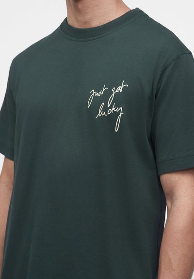 Homme portant un t-shirt vert foncé à manches courtes avec un texte blanc manuscrit "just got lucky" sur le côté gauche de la poitrine.