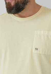 Żółta koszulka z krótkim rękawem, okrągłym dekoltem i kieszonką na lewej piersi. Posiada małe logo na kieszeni, wykonane z gładkiego materiału.