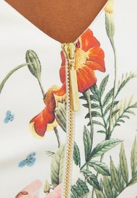 Blommönstrad bodysuit med vit bakgrund, som har röda och gula blommor, gröna blad och en gulddetalj med dragkedja vid halslinningen.