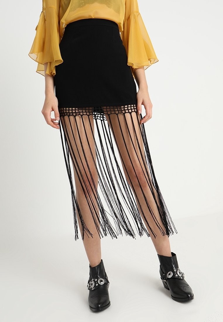 Jupe noire à franges avec une taille ajustée, présentant de longues franges verticales attachées à un haut en filet. Portée avec des bottines noires et un haut jaune.