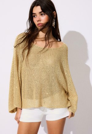 Donna con lunghi capelli scuri che indossa un maglione beige largo a maglia aperta e pantaloncini bianchi, posando su uno sfondo chiaro e semplice.