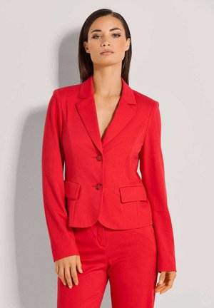 Frau mit glattem Haar, die ein eng anliegendes rotes Blazer und passende Hose trägt, vor einem schlichten hellgrauen Hintergrund stehend.