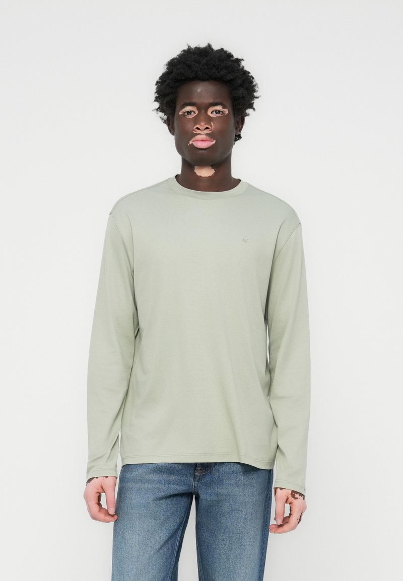 Lys grønn langermet t-skjorte laget av myk bomull. Har rund hals og avslappet passform, kombinert med blå denimjeans.