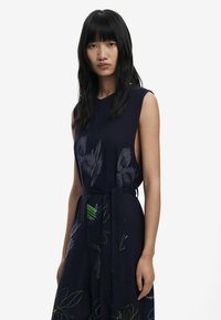 Navy sleeveless dress with a tied waist, featuring vibrant green and gray floral patterns. Smooth fabric with minimal cutout details.