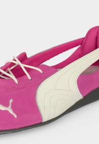 Chaussure plate femme Puma rose et blanche avec lacets blancs et semelle noire, dotée d'une conception à enfiler à l'arrière et d'une texture en daim.