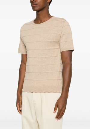 Uomo che indossa un maglione beige a maniche corte texturizzato e pantaloni color crema chiaro, in piedi con postura rilassata su sfondo bianco.