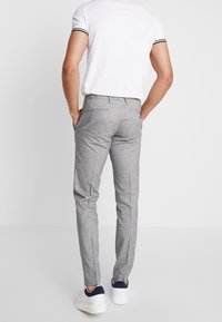 Muž stojící s rukama v kapsách, oblečený v úzkých šedých kalhotách, bílém polo tričku s černým lemem a bílých teniskách s námořnickými akcenty na patách.