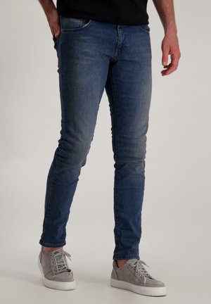 Slim-fit blauwe denim jeans met lichte vervaging, vijf-pocket ontwerp en een middelhoge taille. Gecombineerd met grijze sneakers en een zwart shirt.