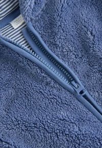 Tissu en polaire bleu avec une texture douce et un détail de fermeture éclair. L'intérieur présente une doublure rayée bleue et blanche.
