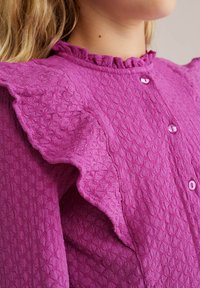 Fuksija teksturirana majica z volanastimi rokavi, z gumbi na sprednji strani in majhnimi roza gumbi. Izkazuje edinstven vzorec tkanine.
