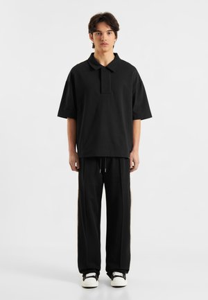 Svart polo skjorte med krage, oversized passform. Kombinert med svarte joggebukser med sidestriper. Enkelt design og myk tekstur.