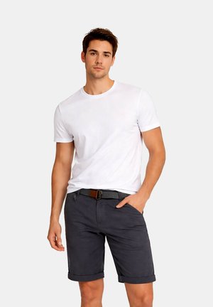 Ung mand iført en ensfarvet hvid kortærmet T-shirt og mørke opkrogede shorts med et vævet bælte, stående med den ene hånd i lommen.
