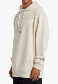Světle béžová mikina s kapucí, s přední kapsou, šňůrkami a vyšitým logem (DC 94). Měkká textura a ribbed manžety a lem.