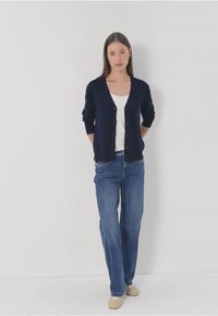 Marinblå kofta med V-ringning, långa ärmar och knappstängning, kombinerad med en vit skjorta och blåjeans. Avslappnad stil, mjuk textur.