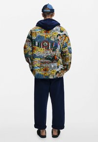 Giacca trapuntata con un design grafico colorato che presenta motivi di animali e paesaggi; il cappuccio blu navy e i pantaloni completano l'outfit.