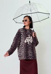 Übergrößenpullover im Leopardenmuster mit einem cremefarbenen Grafik. Kombiniert mit einem kastanienbraunen Rock und hält einen klaren, kuppelförmigen Regenschirm.