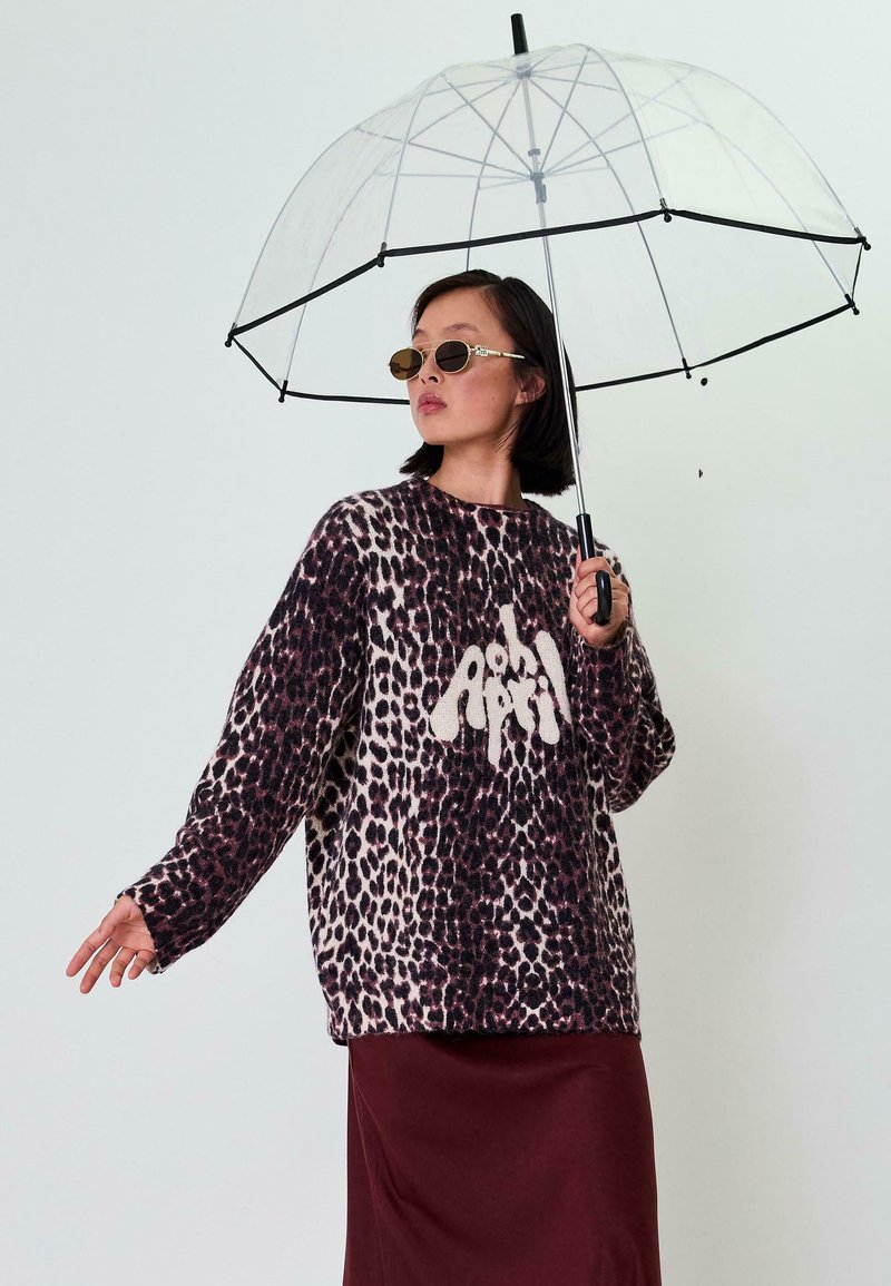 Übergrößenpullover im Leopardenmuster mit einem cremefarbenen Grafik. Kombiniert mit einem kastanienbraunen Rock und hält einen klaren, kuppelförmigen Regenschirm.
