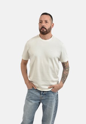 Uomo con braccio destro tatuato che indossa una semplice t-shirt bianca a maniche corte e jeans azzurri, in piedi con le mani nelle tasche.
