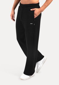 Black sweatpants with a loose fit, elastic waistband, and side pockets. Finished with a small logo detail on the front. Worn with white sneakers.