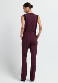 Haut sans manches bordeaux et pantalon bordeaux ajusté, tissu lisse, design sur mesure, poches arrière, et escarpins noirs à bout pointu visibles.