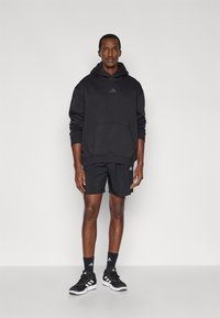 Μαύρο φούτερ με κουκούλα και τσέπη μπροστά, με διακριτικό λογότυπο, σε συνδυασμό με μαύρες βερμούδες και μαύρα αθλητικά παπούτσια με λευκές λεπτομέρειες και κάλτσες.