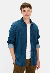 Jeansowa koszula w ciemnym niebieskim kolorze z zapinaniem na guziki, dwoma kieszeniami na klatce piersiowej i podwiniętymi mankietami, noszona na białym T-shircie i brązowych spodniach.