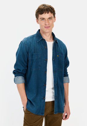 Dunkelblaumjeanshemd mit Knopfleiste, zwei Brusttaschen und hochgerollten Ärmeln, getragen über einem weißen T-Shirt und braunen Hosen.