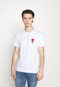 Bílá bavlněná tričko s vyšitou červenou růží na levé části hrudi. Krátké rukávy, kulatý výstřih a volný střih.