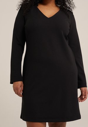 Femme portant une robe noire à manches longues, col en V, longueur au genou, sur un fond neutre.