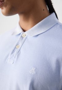 Polo bleu clair en tissu texturé, avec un col blanc et deux boutons, orné d'un petit logo brodé blanc sur la poitrine.