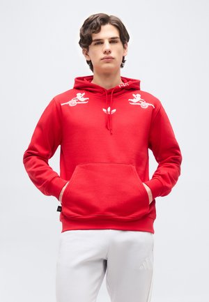 Ung mand iført en rød hættetrøje med hvide engle- og kanongrafikker på skuldrene og hvide bukser, stående med hænderne i forlommerne.