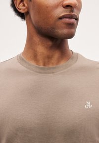 Κοντινό πλάνο του κάτω μέρους του προσώπου και του λαιμού ενός ατόμου που φοράει μπεζ μπλουζάκι με στρογγυλή λαιμόκοψη και μικρό λευκό λογότυπο "M O'P" στο στήθος.