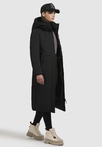 Lange zwarte pufferjas met een capuchon, zijzakken en een getailleerd silhouet. Draagt een crèmekleurige veterschoen met chunky zolen.