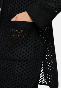 Schwarze gemusterte Oberbekleidung mit einem strukturierten, netzartigen Design. Verfügt über eine Tasche mit glatter Oberfläche, stilvollen Bündchen und einer vorderen Knopfleiste.