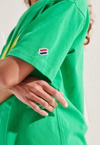 Camiseta de algodón verde de manga corta, con un colorido logo bordado en la manga izquierda. Textura suave y corte cómodo.