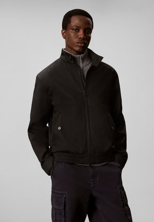Junger Mann trägt eine schwarze Zip-Up-Jacke über einem grauen Rollkragenpullover mit dunklen Cargohosen, steht mit den Händen in den Taschen vor einem schlichten Hintergrund.