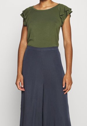Personne portant un haut vert olive à manches courtes avec des manches à volants et un pantalon large taille haute bleu marine foncé.