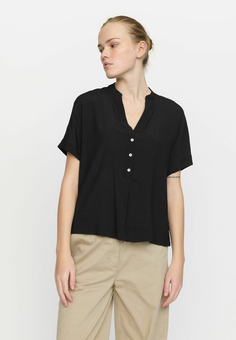 Blusa nera a maniche corte con scollo a V, tessuto strutturato, chiusura frontale con bottoni e vestibilità rilassata. Abbinata a pantaloni beige.