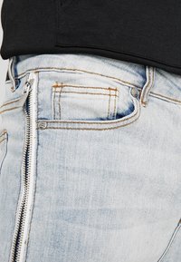 Helle blaue Jeans mit Reißverschluss, die mit einer kleinen Fronttasche und kontrastierenden orangefarbenen Nähten ausgestattet ist.