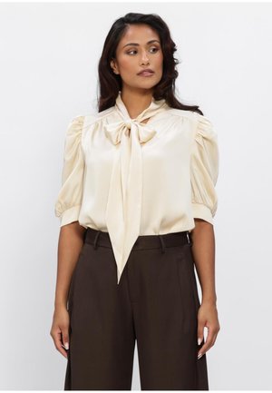 Vrouw die een crèmewitte satijnen blouse draagt met pofmouwen en een strik aan de voorkant, gecombineerd met een hoge taille donkerbruine broek, kijkend naar de zijkant.