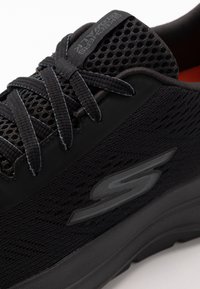 Schwarzer Sportschuh mit Netzpaneelen, gemustertem Textur, gleichen Schnürsenkeln und einem reflektierenden Logo-Element an der Seite. Abgerundete und gepolsterte Sohle.