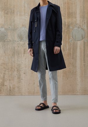 Homme portant un trench-coat bleu marine, une chemise bleu clair, un pantalon gris et des sandales noires, debout contre un mur en béton.
