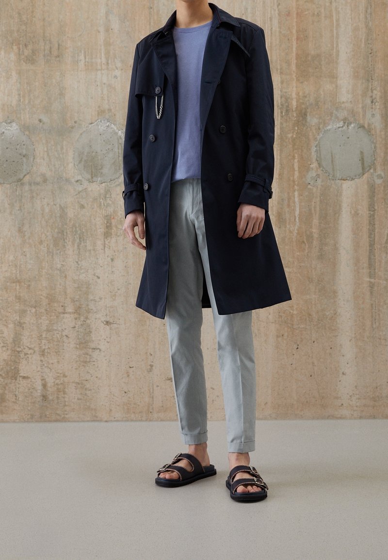 Homme portant un trench-coat bleu marine, une chemise bleu clair, un pantalon gris et des sandales noires, debout contre un mur en béton.